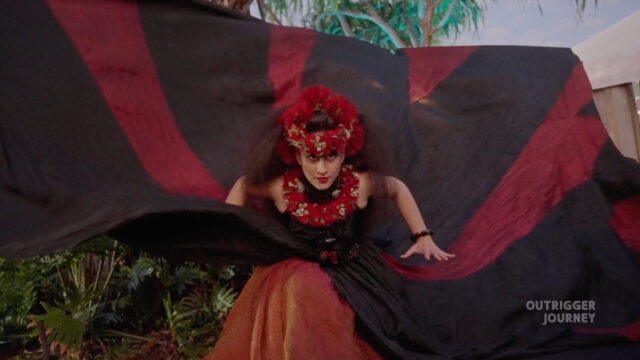 A person in a red and black dress with a floral hair accessory performs a dance with a flowing fabric. The background is lush with greenery and a text overlay reads "OUTRIGGER JOURNEY".