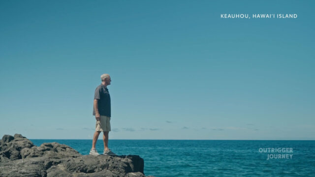 Man standing on a rocky shore gazing out at the calm blue ocean under a clear sky, with text "Keauhou, Hawai'i Island" in the upper right corner and "Outrigger Journey" in the lower right corner.