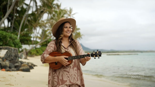 A person wearing a wide-brimmed hat and a patterned shirt playing a ukulele on a sandy beach with palm trees and overcast sky in the background, and calm sea waters extending to the horizon.