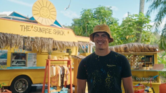 A person wearing a bucket hat and a black t-shirt with colorful paint splatters stands in front of "The Sunrise Shack," a yellow food stand with a thatched roof and signage displaying its name and a stylized sun. Palm trees and tropical foliage are visible in the background.