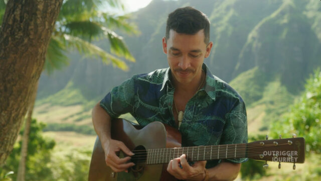 A person wearing a blue patterned shirt is playing an acoustic guitar outdoors with lush greenery and mountains in the background.